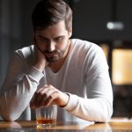 Upset young man drinker alcoholic sitting at bar counter with glass drinking whiskey alone, sad depressed addicted drunk guy having problem suffer from alcohol addiction abuse, alcoholism concept